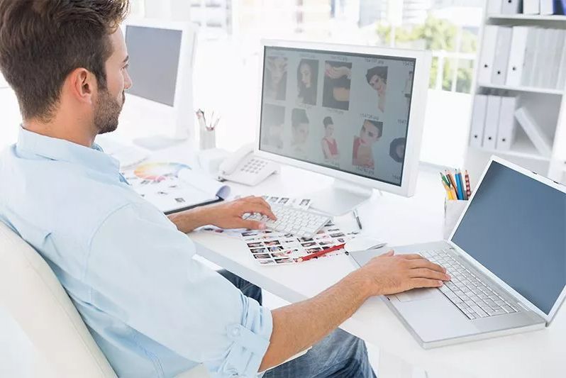 Eine Person arbeitet an einem Computer mit einem zweiten Laptop und grafischen Entwürfen auf dem Schreibtisch