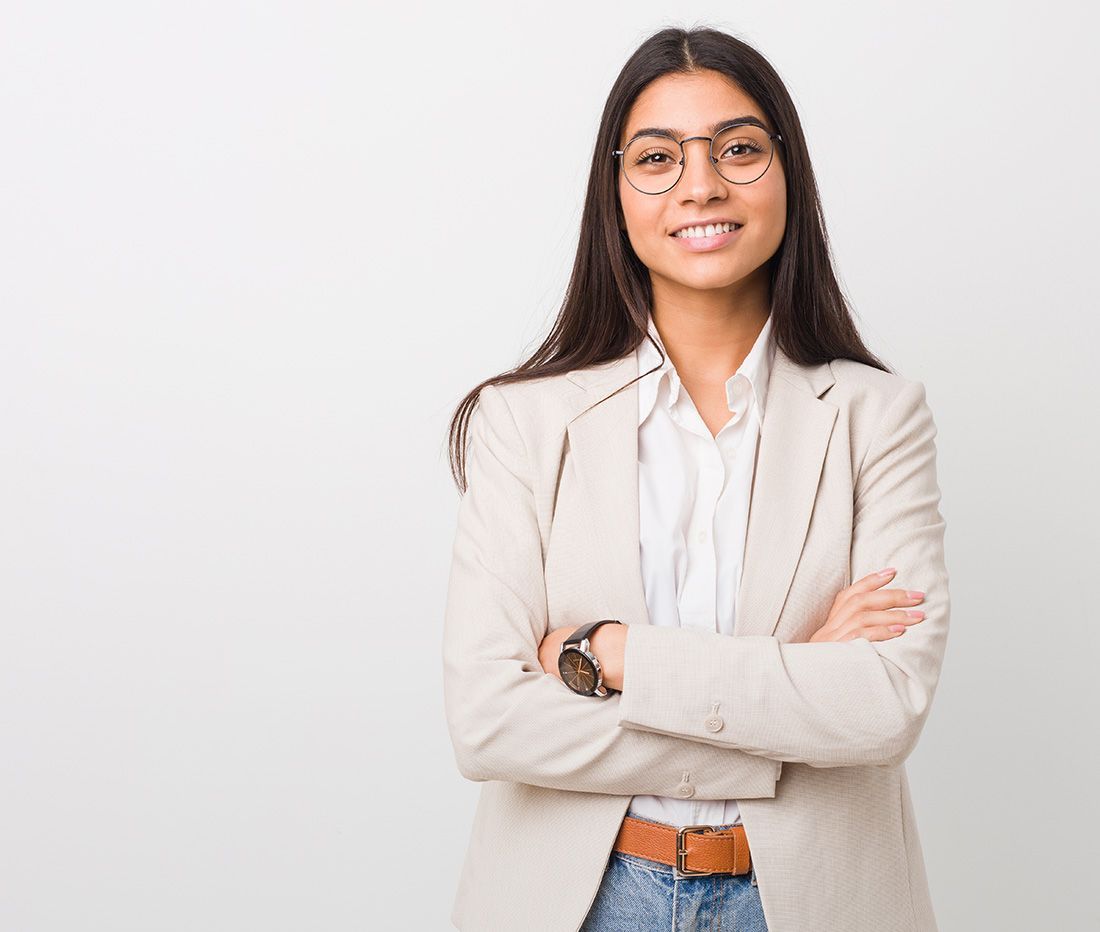 Person in einem beigen Blazer und Jeans mit verschränkten Armen vor einem weißen Hintergrund